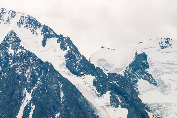 Helicopter in air against background of snowy mountain peak. Rescue mission in glaciers. Aerial tourism in highlands. Atmospheric view on mountain ridge in overcast weather. Icy rock close up.