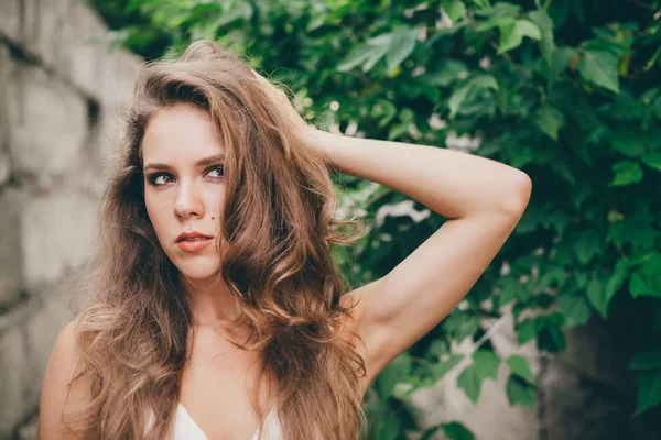 Beautiful mad girl with curly natural hair in white dress near green tree leaves. Summer beauty portrait. Crazy lady in nature outdoors. Alone moody woman with pensive sight in long tunnel in forest.