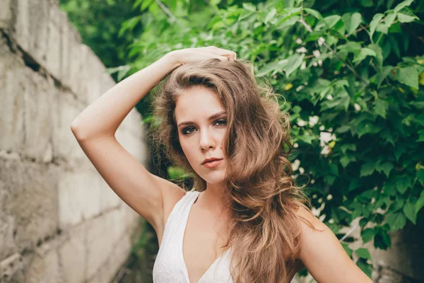 Beautiful moody girl with curly natural hair in white dress near green tree leaves. Summer beauty portrait. Sad lady in nature outdoors. Alone serious woman with pensive sight in long tunnel in forest