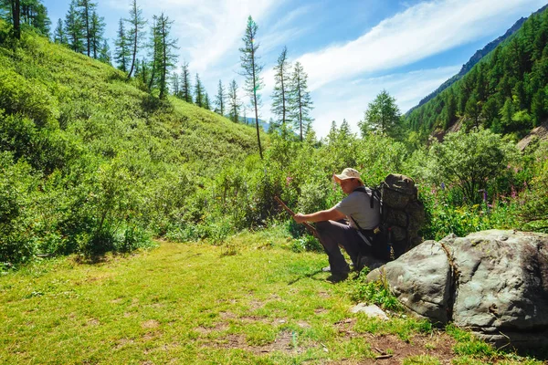 Alone traveler with large backpack and wooden stick. Pilgrim sitting on big stone in valley in sunlight. Hiking in mountains. Pathway among rich vegetation of highlands. Sunny green mountain landscape