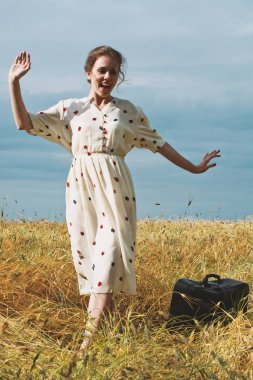 Eski bavulu ile beyaz retro elbiseli neşeli genç kız tatilde altın buğday tarlasında seyahat ediyor. Çavdar arasında Duygu kadın portresi. Güneşli bir günde doğatarafından yolculuk. Seyahatte dans.