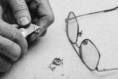 Yaşlı adam kalemi bünyeyle biler. Sanatçının elleri yakın. Gri tonlanınmda gözlüklerin yanındaki masada talaslar. Tek renkli arka plan.