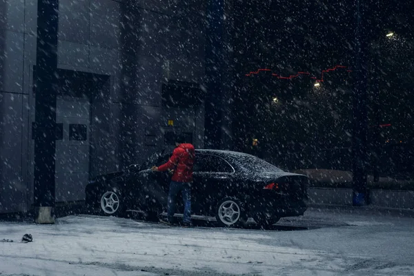 Black car parked near office building in winter evening in strong snow storm. Automobile near auto service in night. Driver near machine near automatic gate for maintenance.