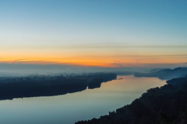 Nehir üzerindeki yüksek kıyıdan manzara. Yoğun sisin altında ormanı olan nehir kıyısı. Şafak suya yansıdı. Pitoresk şafak öncesi gökyüzünde sarı şerit. Görkemli doğanın mistik sakin sabah atmosferik manzara.