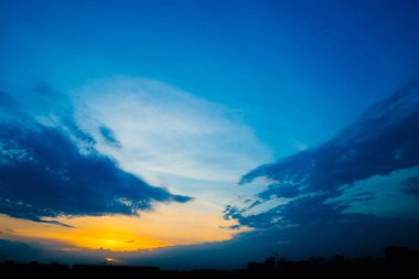 Şehir binalarının siluetleri arkasında atmosferik mavi bulutlu gökyüzü. Kobalt ve kopya alanı için yoğun bulutlar ve parlak sarı güneşli ışık ile gündoğumu turuncu arka plan. Bulutların üzerindeki Siyam cenneti.