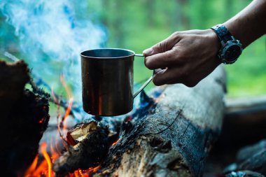 Adam şenlik ateşinde metal kupada çay ısıtıyor. Doğada sıcak içecek. Açık havada çay içmek. İnsan elinde çelik kupa. Gün batımında kamp. Alacakaranlıkta romantik sıcak atmosfer açık . Aktif dinlenme.