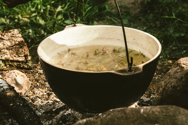 stock image Herbal tea in cauldron is heats up on bonfire, surrounded by stones on background of ashes near green grass. Cooking in open air. Active outdoor recreation. Camping in wild close up.