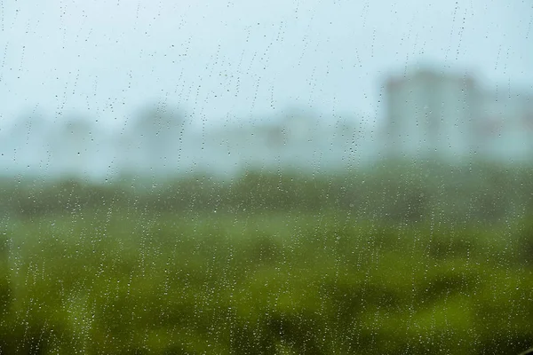 Dirty glass with drops of rain. Raindrops on background from greenery, building and sky in bokeh. City outside window. Droplets and stains close up. Detailed transparent texture in macro.