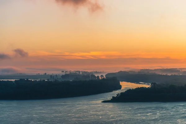 River flows along shore with forest under mist. Channel of river flows around island. Orange glow in dawn sky reflected on water. Colorful morning mystical atmospheric landscape of majestic nature.