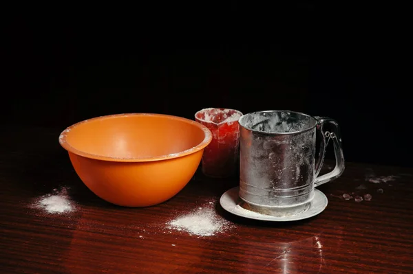 Orange cup, measuring cup, and a steel sieve stand on a wooden table on a black background. Flour.