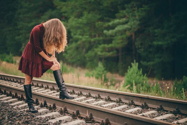 Beautiful sad girl is hiding face by hair. Moody lady in burgundy dress in forest on railway. Depressed lonely girl on railroad at dawn. Sun in curly natural hair in autumn. Bad mood. Offended girl.