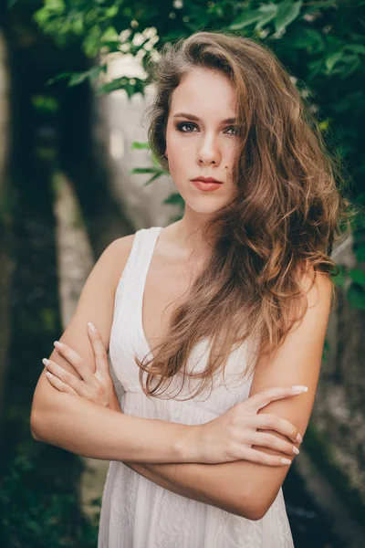 Beautiful moody girl with curly natural hair in white dress near green tree leaves. Summer beauty portrait. Sad lady in nature outdoors. Alone serious woman with pensive sight in long tunnel in forest