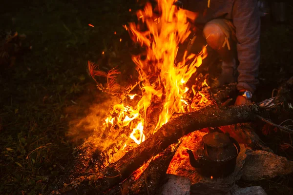 Hot tea in kettle on bonfire. Man bask. Hand throws up brushes in fire. Tea drinking in open air. Active outdoor recreation. Camping in dusk. Atmospheric warm in twilight. Active rest.