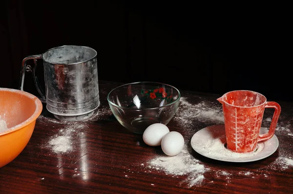 Orange cup, measuring cup, and a steel sieve, two eggs stand on a wooden table on a black background. Flour.