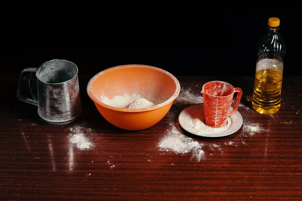 Orange cup, measuring cup, and a steel sieve, sunflower oil stand on a wooden table on a black background. Flour.