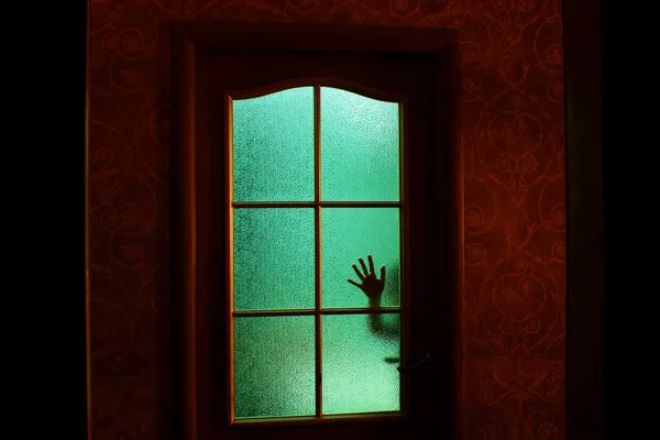 Dark silhouette of hand behind glass in supernatural green light. Locked alone in room behind door on Halloween. Nightmare of child with aliens, monsters and ghosts. Evil in home. Inside haunted house