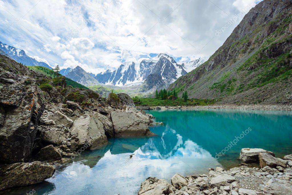Mountain lake is surrounded by large stones and boulders on front of giant beautiful glacier. Amazing snowy mountains. Ridge with snow. Wonderful atmospheric landscape of majestic nature of highlands.