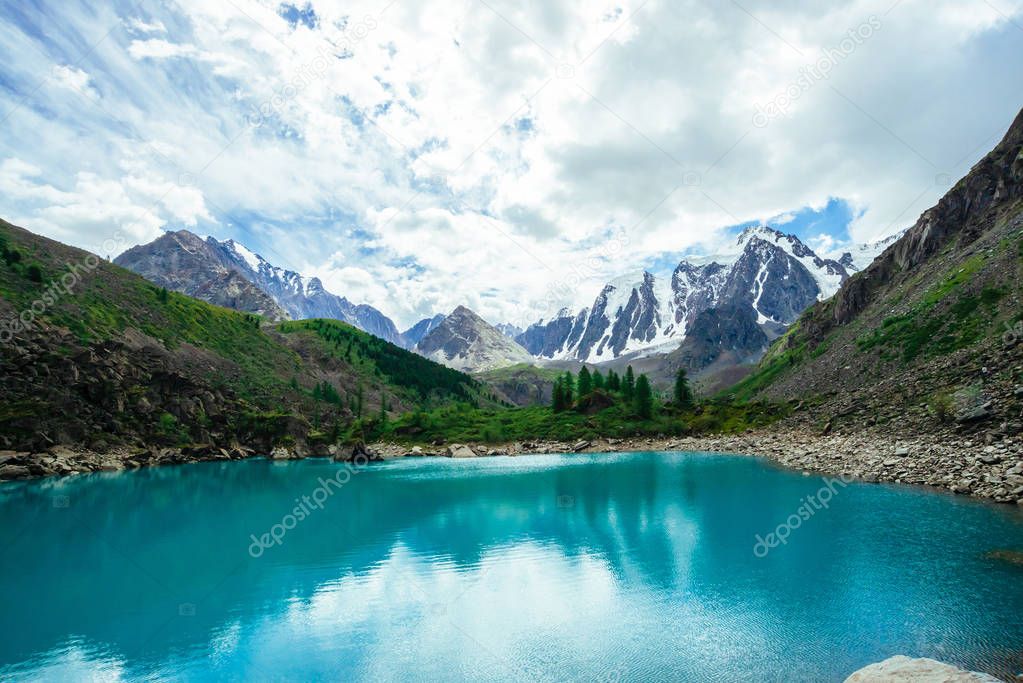 Mountain lake is surrounded by large stones and boulders on front of giant beautiful glacier. Amazing mountain in shape of pyramid. Snowy ridge under cloudy sky. Wonderful atmospheric landscape.