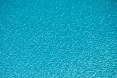 Sakin masmavi temiz su yüzeyinin inanılmaz dokulu arka planı. Dağ gölünde güneş ışığı yakın. Güneşli bir günde parlak suda güzel dalgalar. Harika rahatlayın doku.