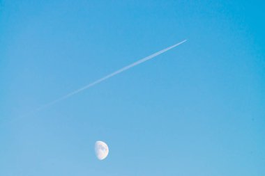 Berrak mavi gün gökyüzünde ay üzerinde jet yoğuşma izi. Minimalist mavi arka plan. Uçak çapraz olarak uçar. Uçak hava sahasında uçuyor. Gökyüzündeki beyaz ay.