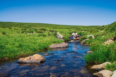 Dağ Deresi'nde taş üzerinde kameralı gezgin. Turist macerası. Dağlarda yürüyüş. Yaylaların zengin bitki örtüsü. Deredeki berrak su akıntısı. Görkemli doğanın canlı güneşli manzara.