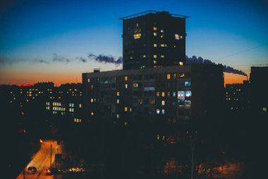 Sıcak gün batımı gökyüzünün arka planında aydınlık pencerelere sahip inanılmaz bina siluetleri. Şafakta şehrin üstündeki borudan duman. Alacakaranlıkta parlayan pencereler. Minimalist şehir manzarası. Harika kentsel gündoğumu.