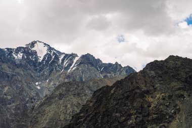 Bulutlu gökyüzünün altındaki dev kayalık sırtta kar. Atmosferik karanlık dağ yamacı. Bulutlu havalarda inanılmaz karlı dağ sırası. Harika kayalar. Yaylaların görkemli doğasının harika gölgeli manzarası
