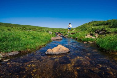 Dağ Deresi'nde taş üzerinde kameralı gezgin. Turist macerası. Dağlarda yürüyüş. Yaylaların zengin bitki örtüsü. Deredeki berrak su akıntısı. Görkemli doğanın canlı güneşli manzara.