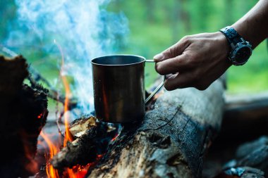 Adam şenlik ateşinde metal kupada çay ısıtıyor. Doğada sıcak içecek. Açık havada çay içmek. İnsan elinde çelik kupa. Gün batımında kamp. Alacakaranlıkta romantik sıcak atmosfer açık . Aktif dinlenme.