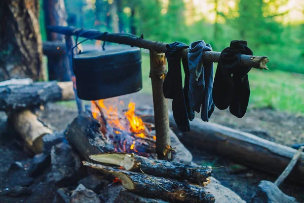 Drying wet clothing on the bonfire during camping. Socks drying on fire. Cauldron and kettle above campfire. Cooking of food on nature. Firewood and branches in fire. Active rest in forest.