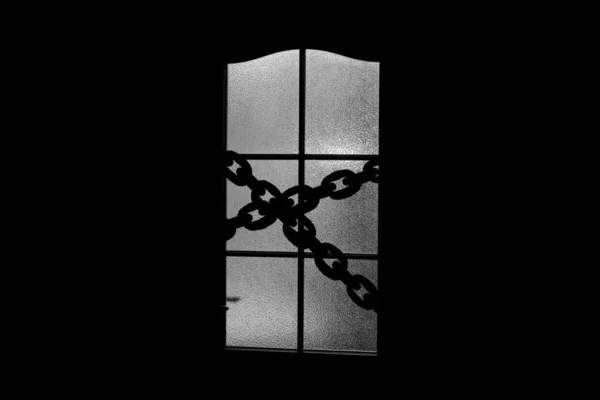 Dark silhouette of glass door with chain. Locked on chain alone in room behind door on Halloween. Evil in home. Inside haunted house in monochrome. Alone in dark. Night kidnapping in grayscale.