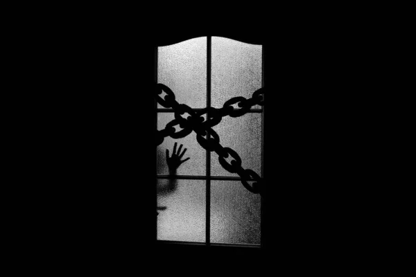 Dark silhouette of hand behind glass door with chain. Locked alone in room behind door on chain on Halloween. Night kidnapping. Evil in home on monochrome. hild inside haunted house in grayscale.