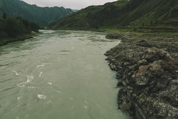 Gloomy mountain landscape with road along river on opposite bank. Stony bank of river. Dark green color of water. Eerie atmosphere in overcast rainy weather in cinematic faded tones. Horror style.