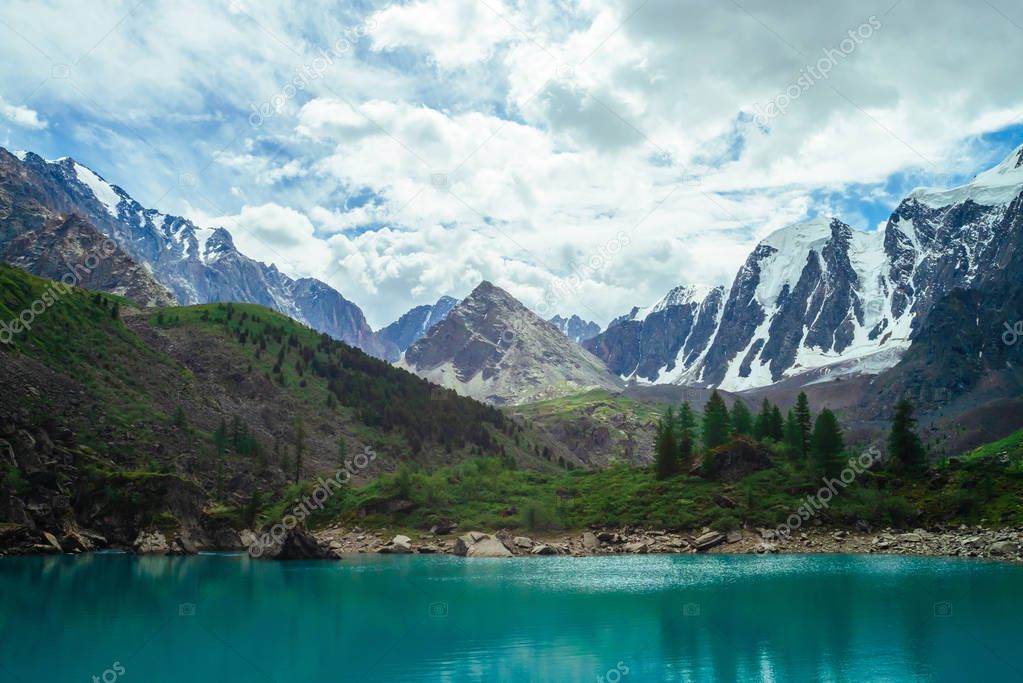 Mountain lake on front of giant beautiful glacier. Amazing mountain in shape of pyramid. Snowy ridge under cloudy sky. Wonderful atmospheric landscape of majestic nature. Coniferous trees in highlands