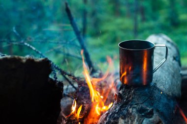 Metal kupadaki çay şenlik ateşinde taşüzerinde ısınır. Doğada sıcak içecek. Açık havada çay içmek. Gün batımında kamp. Alacakaranlıkta romantik sıcak atmosfer açık . Aktif dinlenme.