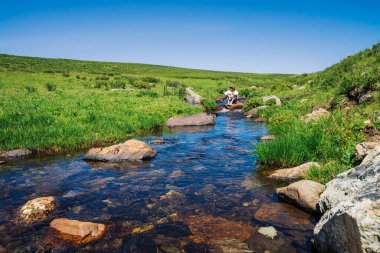 Dağ Deresi'nde taş üzerinde kameralı gezgin. Turist macerası. Dağlarda yürüyüş. Yaylaların zengin bitki örtüsü. Deredeki berrak su akıntısı. Görkemli doğanın canlı güneşli manzara.