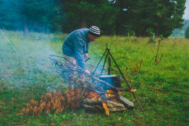Turist ateşi yakSa. Gezgin kampta ateş yakıyordu. Adam kamp ateşine kuru dal koyar. Aktif açık hava rekreasyon. Vahşi doğaya. Gün batımında kamp. Alacakaranlıkta atmosferik sıcak. Aktif dinlenme.