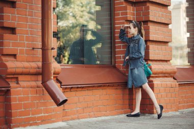 Mavi denim elbiseli genç kız kırmızı (kahverengi) tuğla açık bina penceresinde bakmak. Dükkanın yakınında genç ciddi çocuk piddle. Büyük pencerede hayalperest refleksi. Kız alışveriş yapmak istiyor.