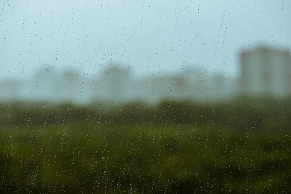 Dirty glass with drops of rain. Raindrops on background from greenery, building and sky in bokeh. City outside window. Droplets and stains close up. Detailed transparent texture in macro.