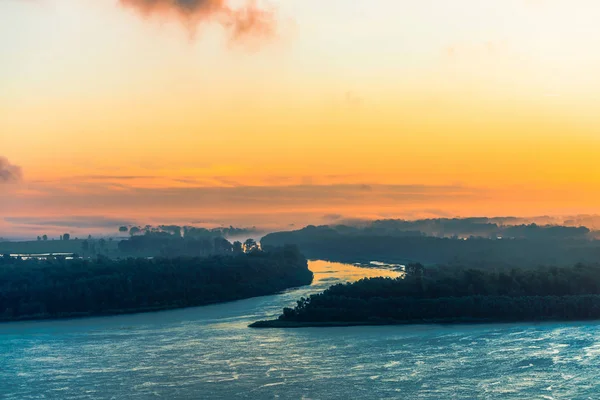 Azure river flows along shore with forest under mist. Channel of river flows around island. Orange glow in dawn sky. Colorful morning mystical atmospheric landscape of majestic nature.