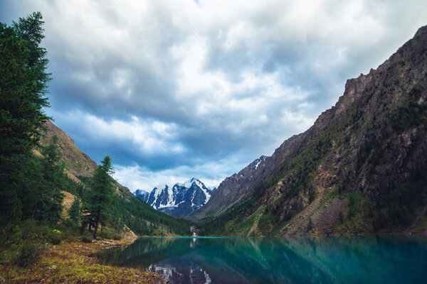 Wonderful mountain lake with view on giant glacier. Amazing huge mountains with conifer forest. Larch tree on water edge. Morning landscape of majestic nature of highlands. Cloudy mountainscape.