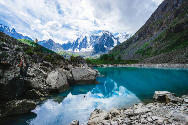 Mountain lake is surrounded by large stones and boulders on front of giant beautiful glacier. Amazing snowy mountains. Ridge with snow. Wonderful atmospheric landscape of majestic nature of highlands.