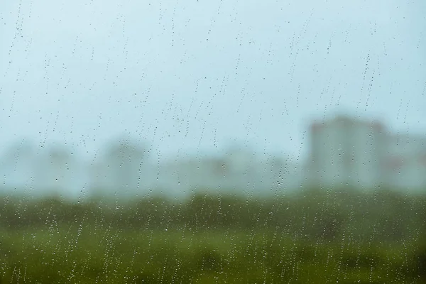 Dirty glass with drops of rain. Raindrops on background from greenery, building and sky in bokeh. City outside window. Droplets and stains close up. Detailed transparent texture in macro.