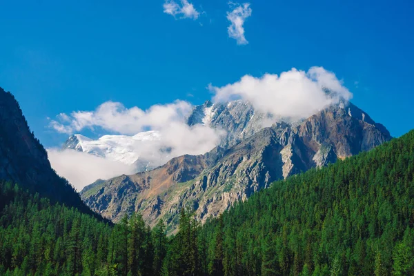 Giant rock in sunny day. Rocky ridge with snow behind hills with conifer forest cover. Clouds on top of huge snowy mountain range under blue sky. Atmospheric highland landscape of majestic nature.