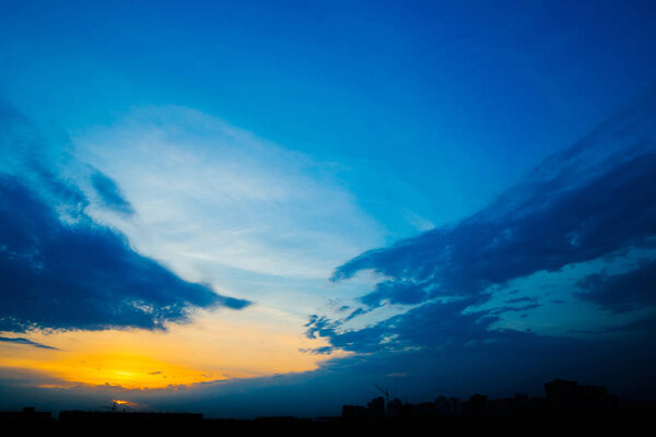 Atmospheric blue cloudy sky behind silhouettes of city buildings. Cobalt and orange background of sunrise with dense clouds and bright yellow sunny light for copy space. Cyan heaven above clouds.