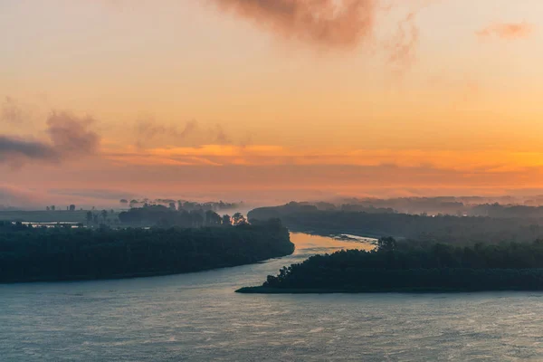 River flows along shore with forest under fog. Channel of river flows around island. Orange glow in dawn sky reflected on water. Colorful morning mystical atmospheric landscape of majestic nature.