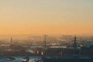 Şafakta şehirde elektrik hatları. Gün doğumunda sis arasında kentsel binaların siluetleri. Sıcak turuncu sarı gökyüzünde yüksek voltaj kabloları. Gün batımında güç endüstrisi. Şehir güç kaynağı. Sis kentsel arka plan.