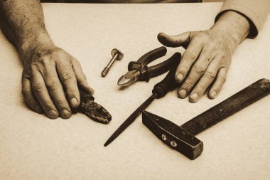 Araçları (nippers, çekiç, pliers, ahududu, cıvata) ve sepya tonları yaşlı çalışan adamın elleri ile tablo kompozisyonu. Tek renkli arka plan.