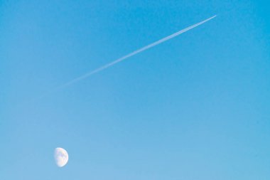 Berrak mavi gün gökyüzünde ay üzerinde jet yoğuşma izi. Minimalist mavi arka plan. Uçak çapraz olarak uçar. Uçak hava sahasında uçuyor. Gökyüzündeki beyaz ay.