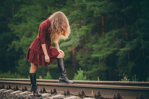 Beautiful sad girl is hiding face by hair. Moody lady in burgundy dress in forest on railway. Depressed lonely girl on railroad at dawn. Sun in curly natural hair in autumn. Bad mood. Offended girl.